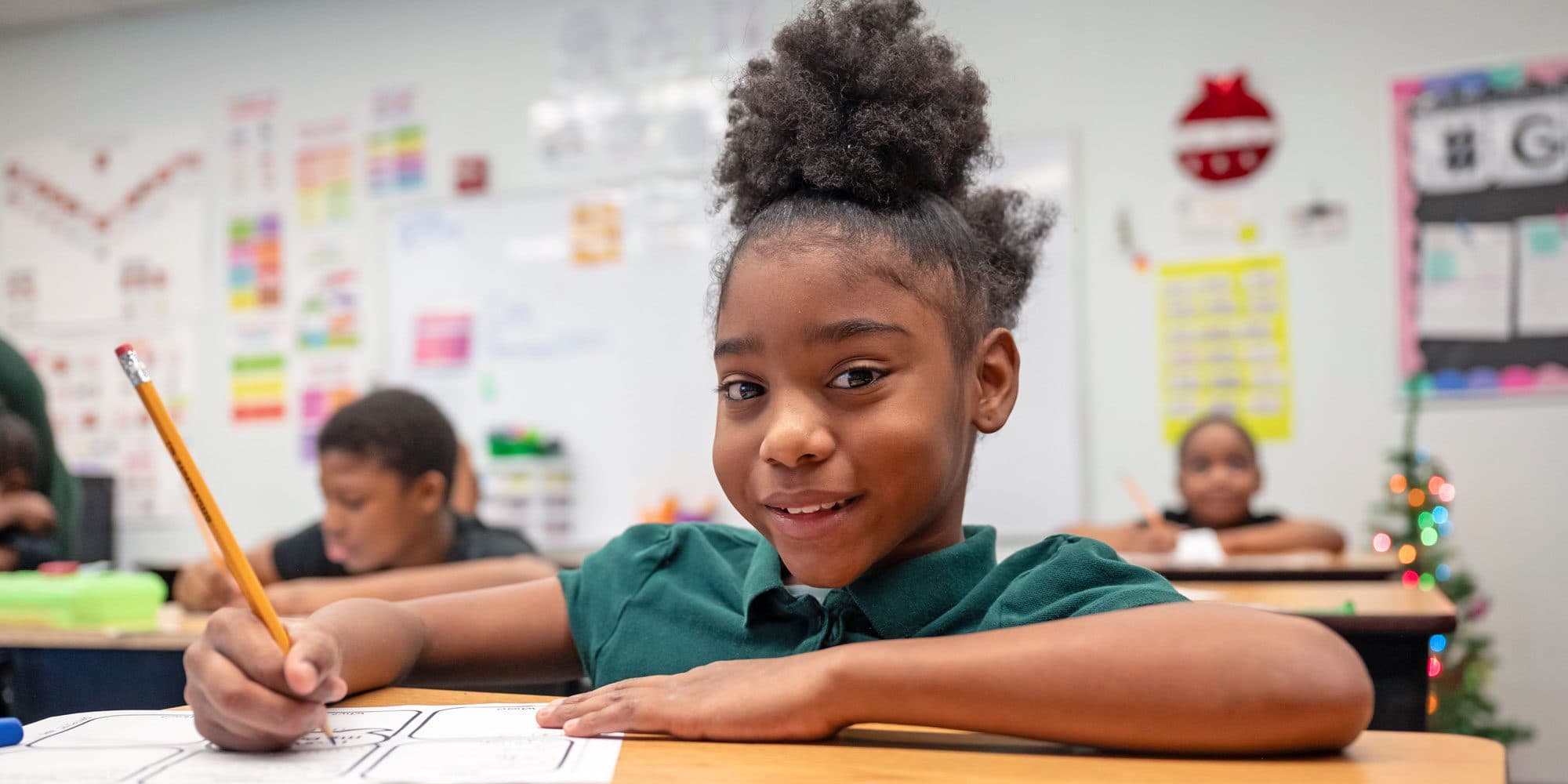 Smiling student writing with a pencil at a classroom desk.